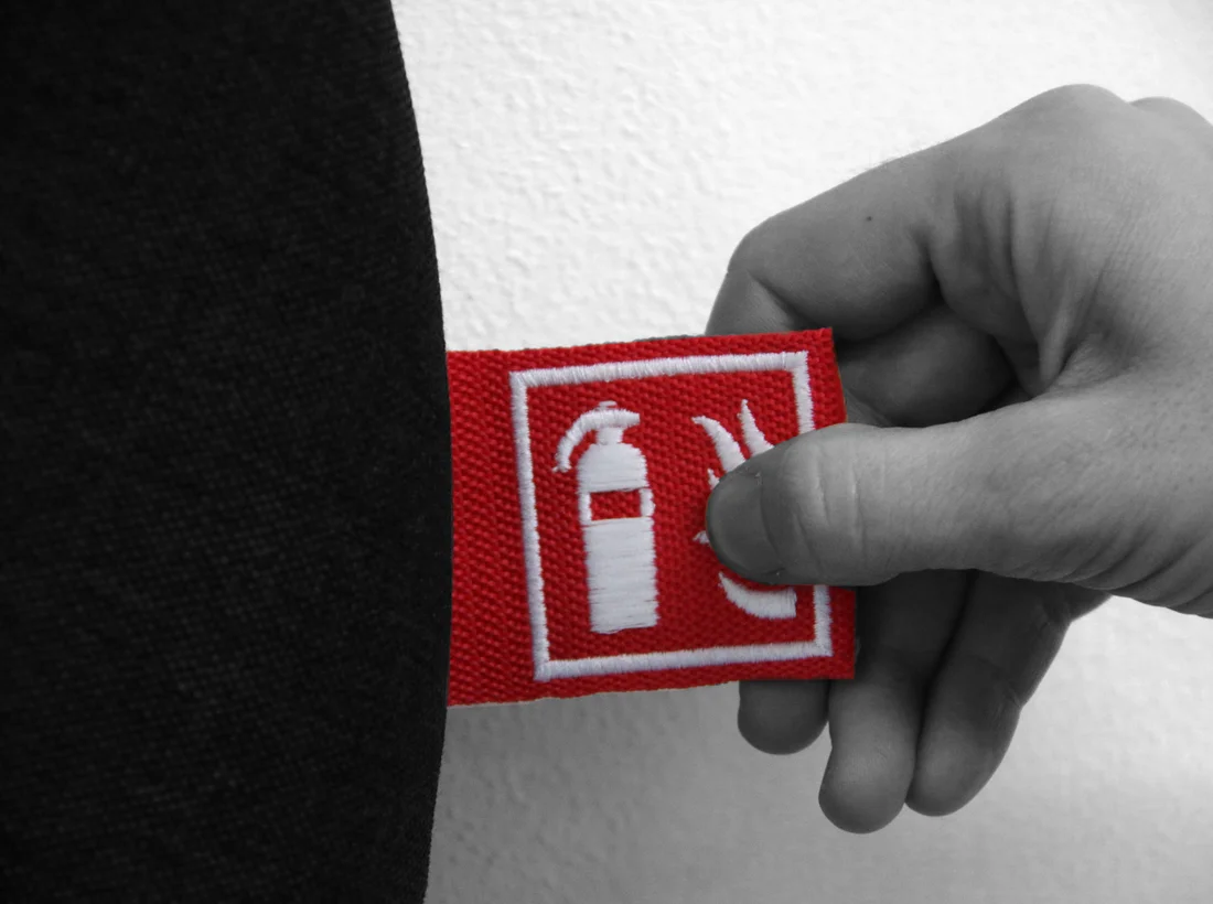 A hand holding a fire-safety badge — the international EN 3-7 extinguisher icon — against a sleeve. Red on black-and-white editorial.